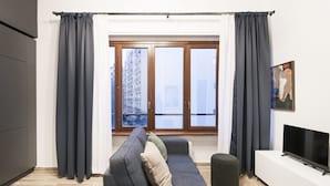 Uma sala de estar moderna com um sofá cinza, um banquinho preto, uma TV de tela plana e uma janela grande com cortinas.