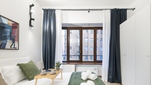 Quarto de hotel moderno com uma cama, uma mesinha com uma chávena e um livro, um cobertor verde e vista para edifícios pela janela.