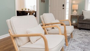 A room with two wooden chairs with white cushions, a grey patterned rug, a grey sofa, a small table with a lamp, and a door with a 'HELLO' sign. - Pet Friendly 3BR | Fenced Yard + Near RAFB (Warner Robins)