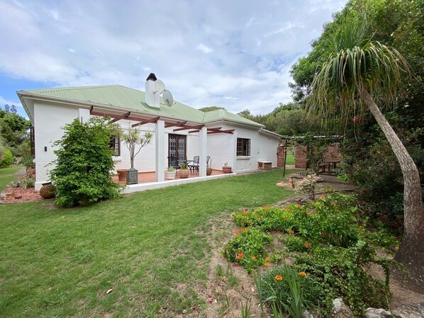A white house with a green roof, a covered patio, and a garden with various plants and flowers. - Beautiful, charming property for 6 to 8 people. Pool. Approx. 400m to the golf course (Port Alfred)
