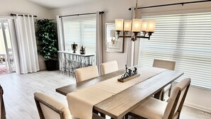 Een eetgedeelte met een houten tafel, beiges bekleden stoelen, een geweven placemat en een kroonluchter.