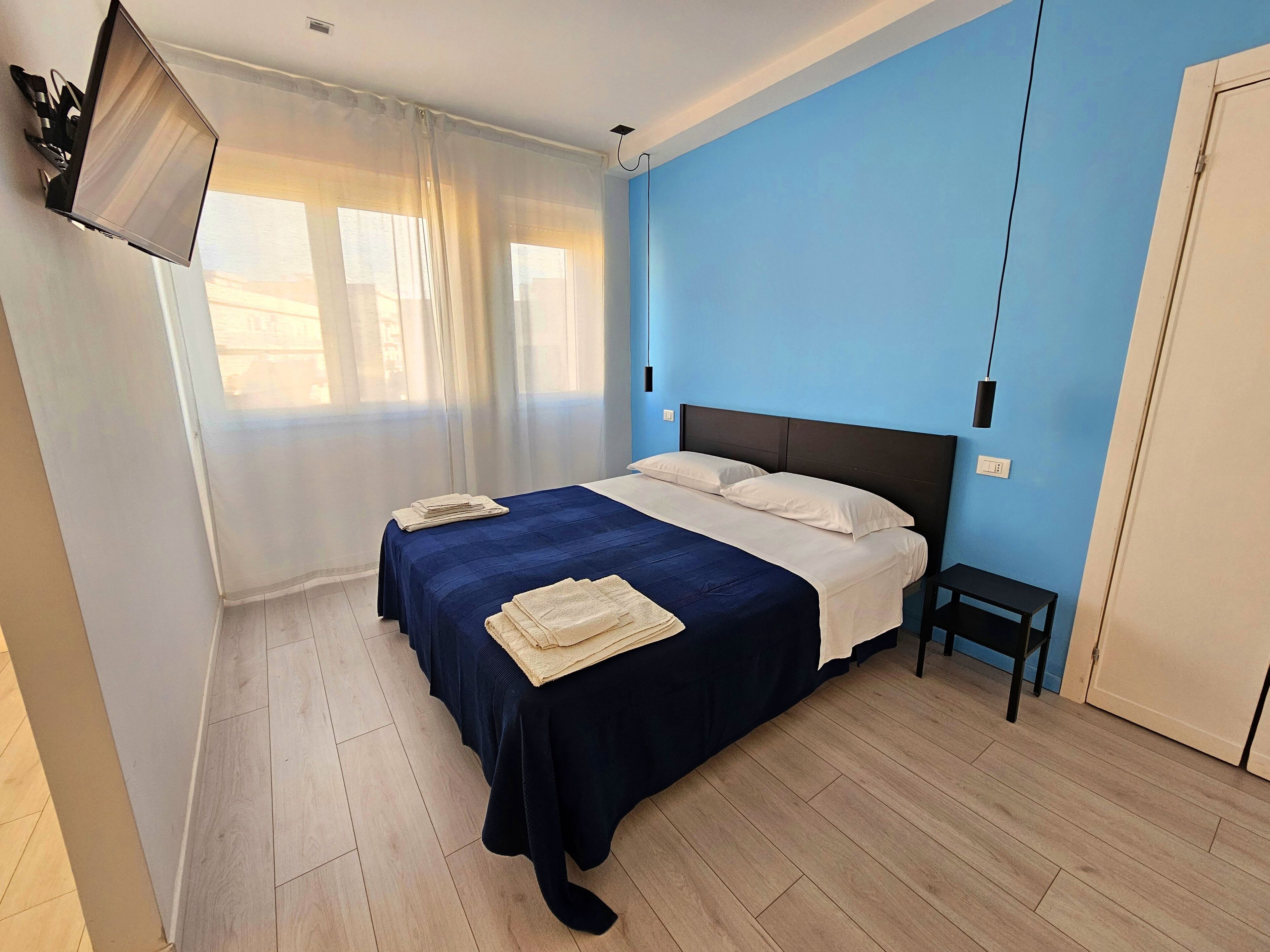 Un dormitorio con cama, mesita de noche, televisor montado en la pared y ventana con cortinas.