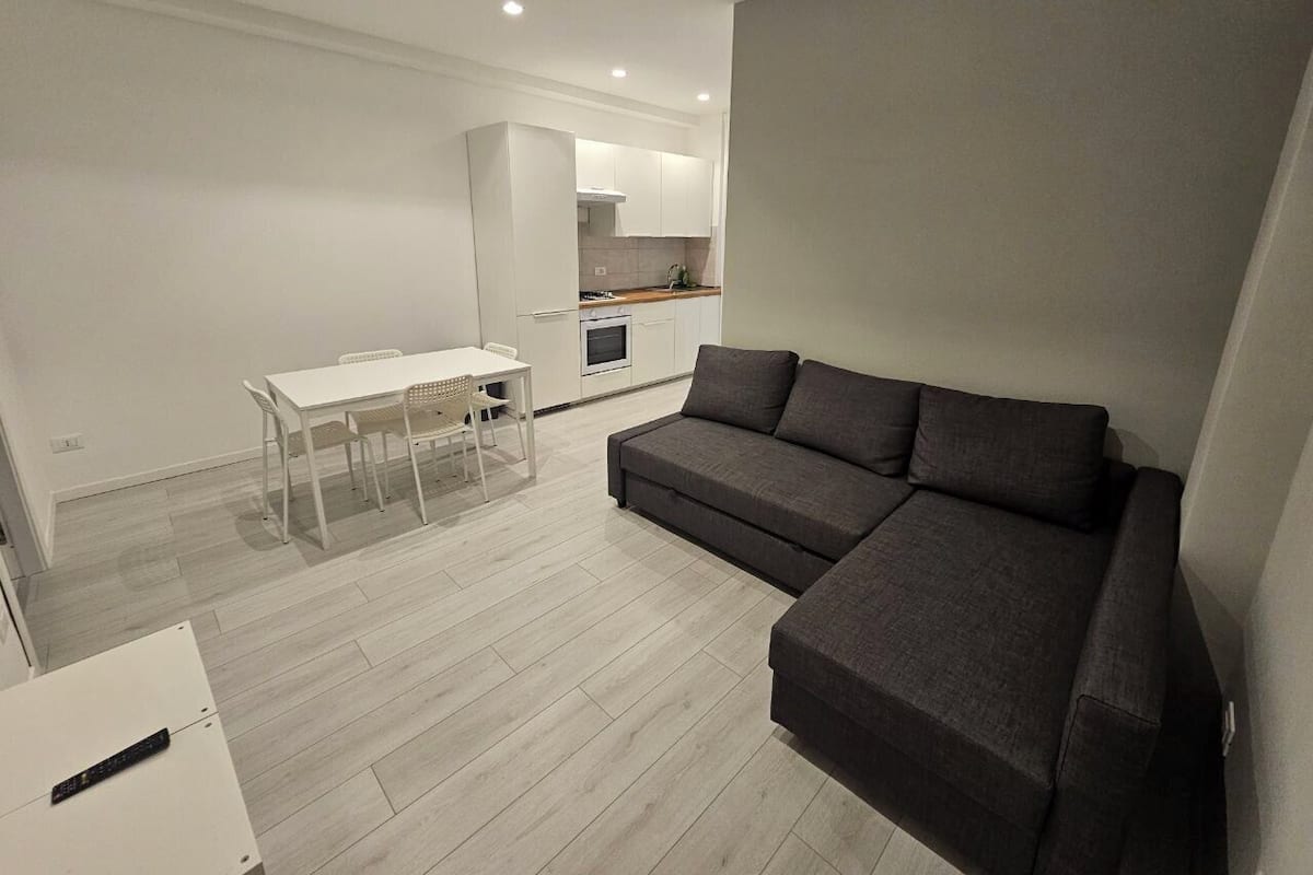 Un salon moderne avec un canapé gris, une table à manger avec des chaises et, au fond, un coin cuisine.