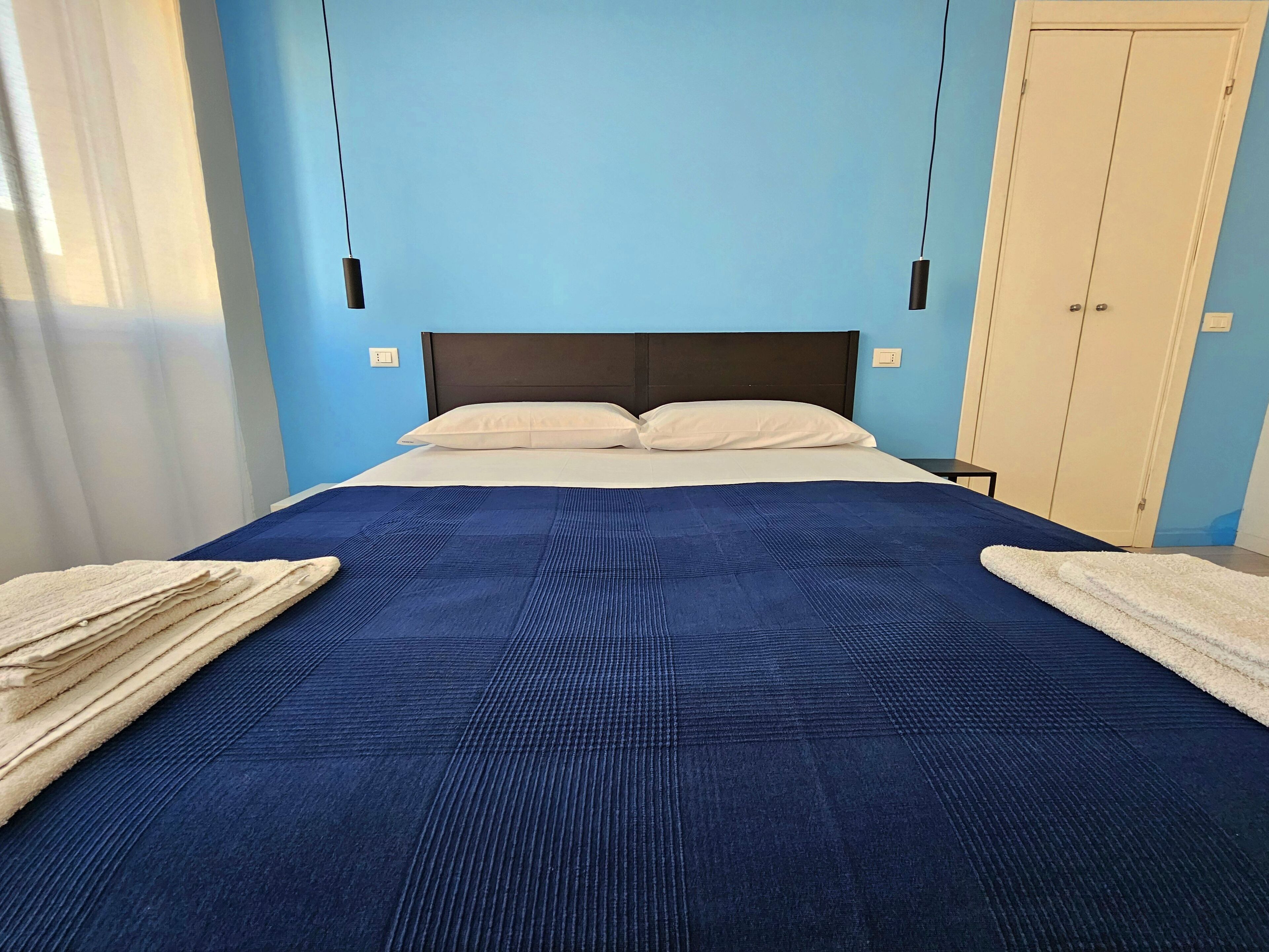 Un letto ben rifatto con copriletto blu e due cuscini, un comodino e un armadio in una stanza dalle pareti azzurre.