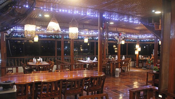 Un restaurant avec des tables et des chaises en bois, des luminaires suspendus et une vue sur le ciel nocturne grâce à de grandes fenêtres.