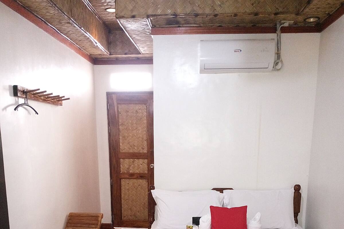 Quarto de hotel com cama, um banquinho de madeira, uma porta e um ar condicionado fixado na parede.