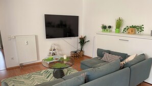 A modern living room with a grey sofa, a glass coffee table with leaf designs, a flat-screen TV mounted on the wall, and a small shelf with potted plants. - Living by the historic town hall (Michelstadt)