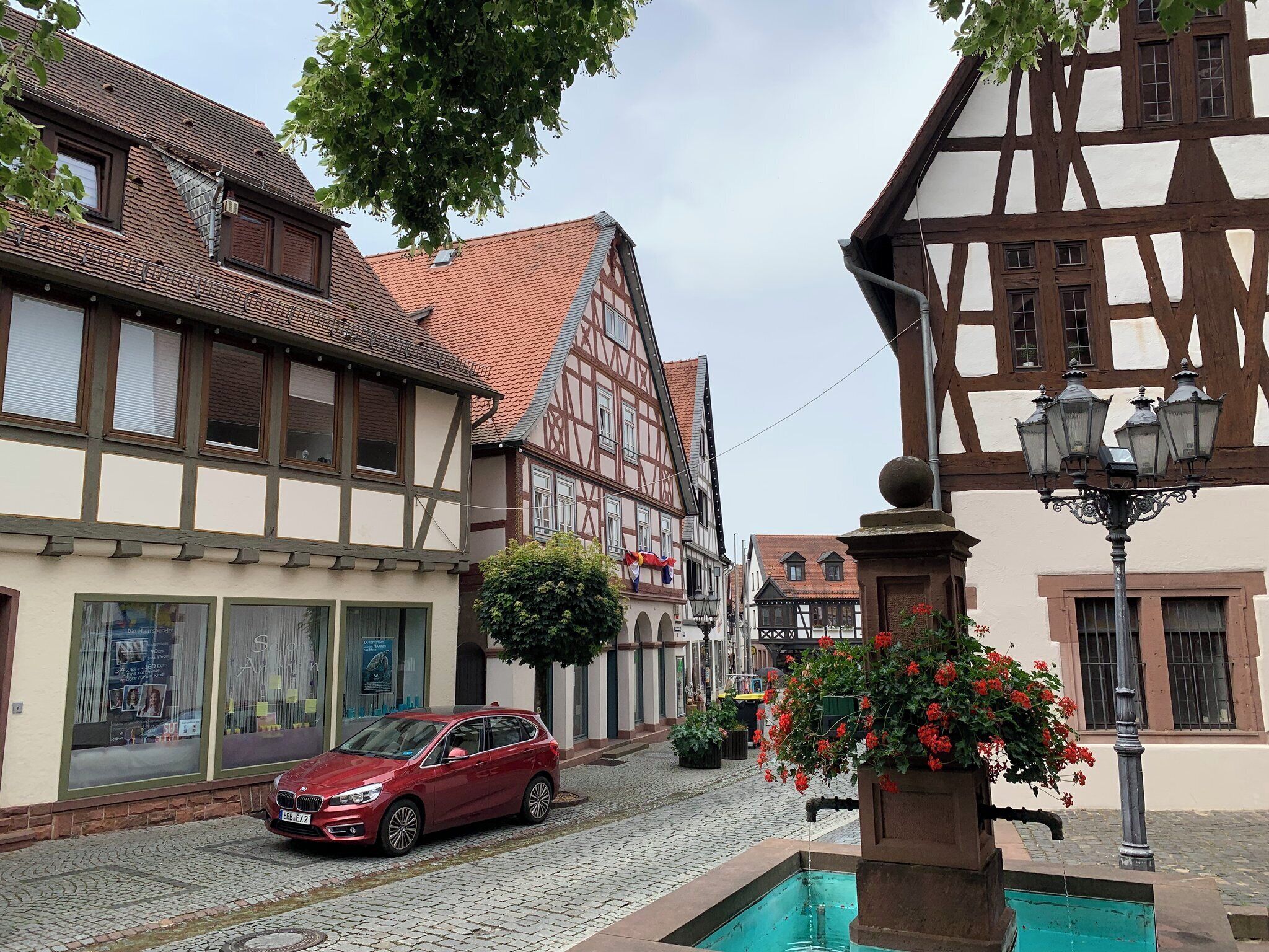 Een rode auto geparkeerd op een geplaveid straatje in een traditioneel Europees stadje met vakwerkhuizen en een fontein.