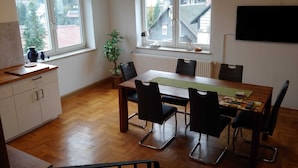 A modern kitchen with a dining area, wooden flooring, white cabinets, and large windows with a view of buildings. - Quiet vacation home with lots of space (Masserberg)