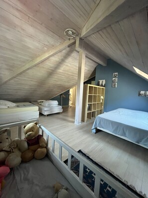 Ein Zimmer mit schrägem Dach, einem Bett, einem Babybett, einem Teddybären und einem Bücherregal.