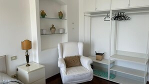 A bedroom with a white bed, a nightstand with a lamp, a chair, a wardrobe with shelves, and a small shelf with decorative items. - Sea view apartment in the city center, 3 minutes from the station and port (Salerno)