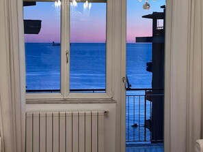 Uitzicht vanuit een raam op een wateroppervlak tijdens zonsondergang, met een balkon en hekwerk.