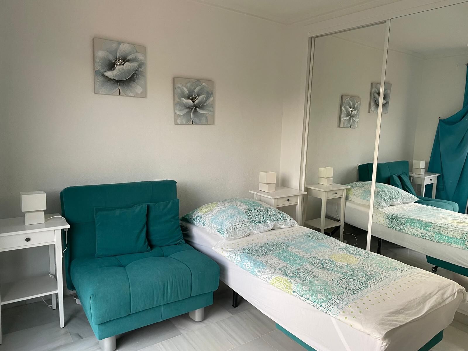 Une chambre avec un canapé de couleur turquoise, deux lits simples, une table de chevet avec une lampe et un grand miroir.
