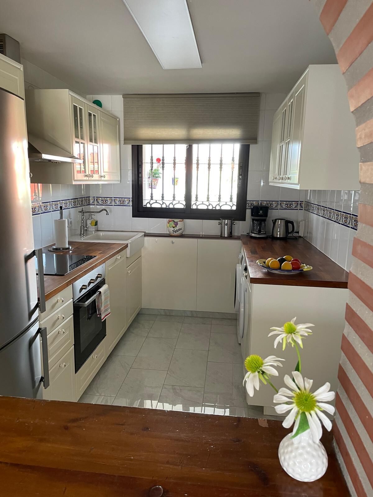 A kitchen with white cabinets, a stainless steel refrigerator, a built-in oven, and a window with a view of the street.