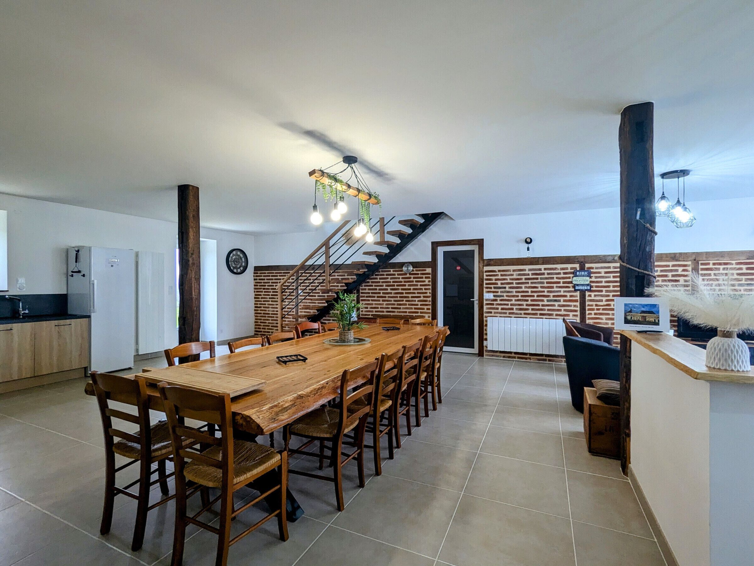 A spacious dining area with a long wooden table and chairs, a kitchen area with white appliances, and a brick wall with a staircase leading to an upper level.