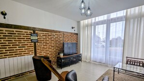 Een woonkamer met een bakstenen muur, een televisie, een houten stoel, een glazen tafel en een groot raam met gordijnen.