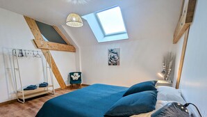 Slaapkamer met schuine balken, dakraam, bed met blauw beddengoed, houten stoel, klein tafeltje met lamp en een kapstok aan de muur.