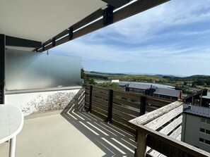 A balcony with a view of a lake and hills, a table, and a chair.