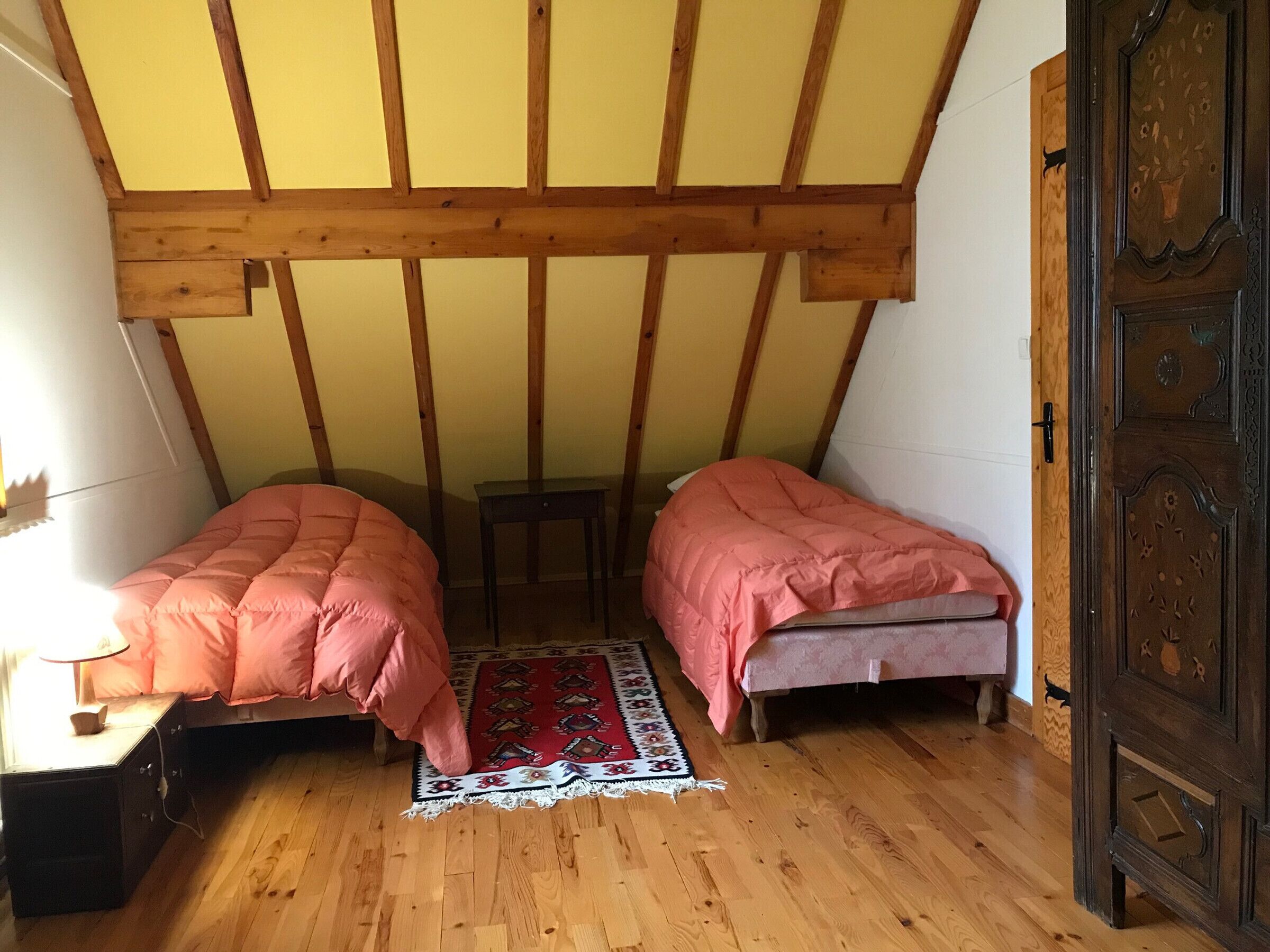 A small, cozy room with two single beds, a wooden floor, a red patterned rug, and a wooden ceiling structure.