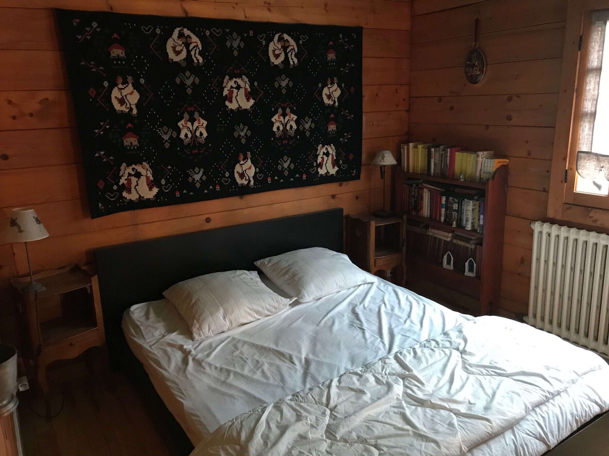 A bedroom with a bed, a nightstand, a bookshelf, and a tapestry on the wall.