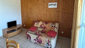 Una habitación con pared de paneles de madera, un sofá con obra de arte tradicional, un televisor sobre un mueble de madera y una ventana con cortinas.