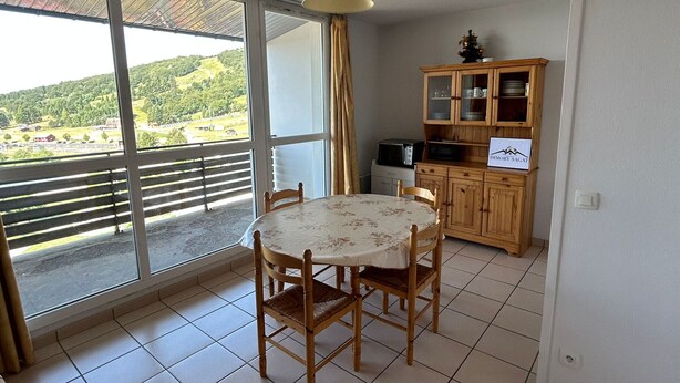 Un comedor con mesa y sillas, una cocina con armarios, y un amplio ventanal corredero que ofrece vistas a un paisaje verde.