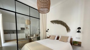A modern bedroom with a large bed, a glass partition separating it from a kitchen, and a decorative wall piece.
