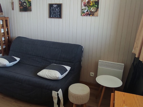 Un sofá de color oscuro con dos almohadas, un taburete redondo pequeño, una mesa blanca y un marco de pared con la palabra 'Juice'.
