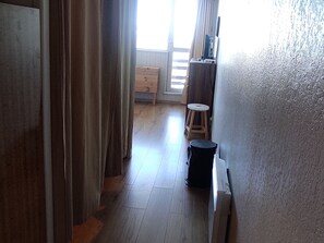 A hallway with a wooden floor, a small stool, a trash bin, and a window with curtains.