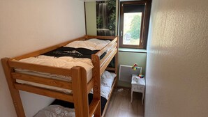 Une petite chambre avec un lit simple et un lit superposé en bois, une fenêtre donnant sur la verdure et une petite table de chevet avec une lampe.
