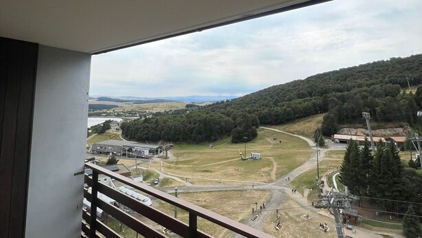 A balcony with a view of a hilly landscape, a road, and a building with a sign. - Apartment at the slopes with private garage (Besse-et-Saint-Anastaise-Super Besse)