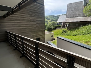 A balcony with a wooden railing overlooking a green landscape and a building with a sloped roof. - Cozy 1-bedroom apartment in Super Besse center (Besse-et-Saint-Anastaise-Super Besse)