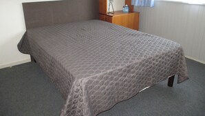 A single bed with a quilted bedspread in a room with a wooden headboard, a nightstand with a lamp, and a window with a blue curtain.