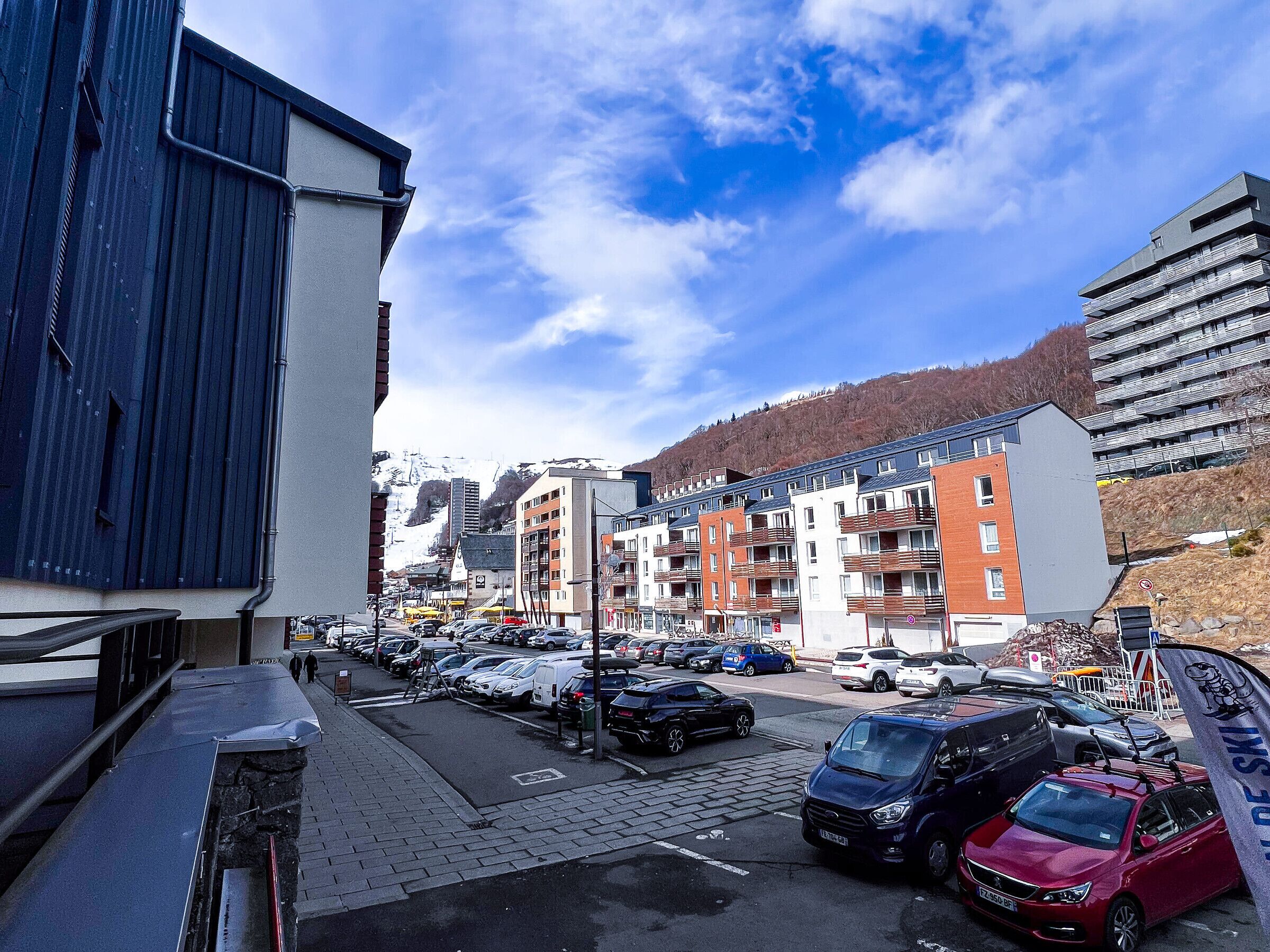 Un parking rempli de voitures, un immeuble avec des balcons et une montagne en arrière-plan.
