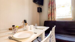 A dining area with a table set for two, a television mounted on the wall, and a window with curtains.