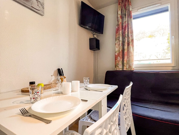 A dining area with a table set for two, a television mounted on the wall, and a window with curtains.