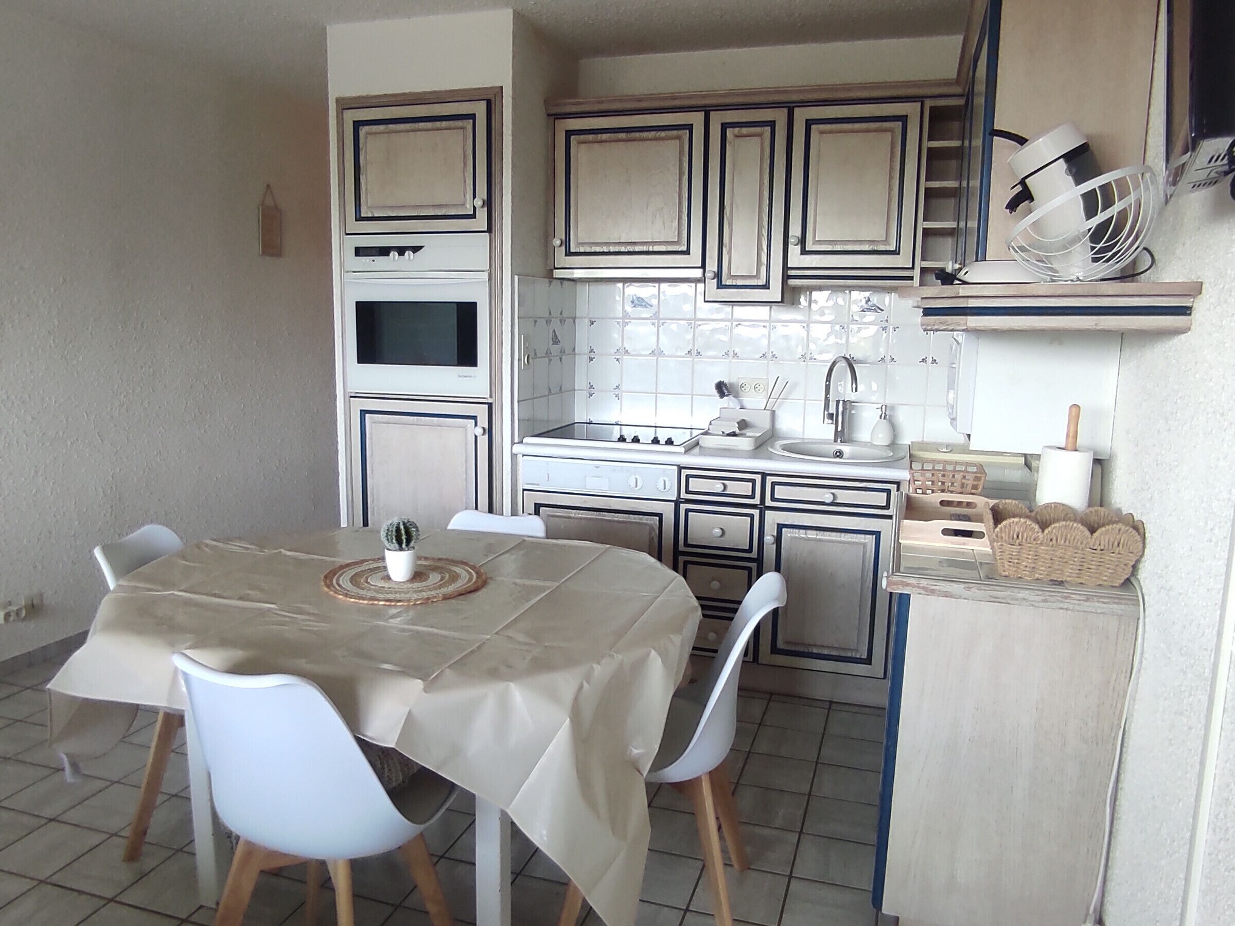Una cocina compacta con una mesa de comedor, sillas, una pequeña planta en maceta y un ventilador blanco en un estante.