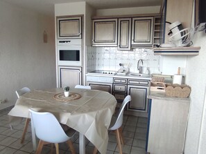 A compact kitchen with a dining table, chairs, a small potted plant, and a white fan on a shelf.
