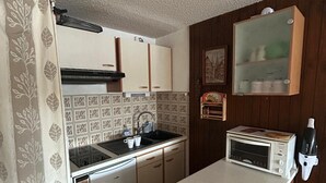 A compact kitchen with a sink, stove, and microwave. There is a wooden cabinet above the sink and a small shelf with items on the wall. - Studio cabin 4 pers, center, WIFI (Besse-et-Saint-Anastaise-Super Besse)