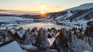 Schneebedeckte Landschaft mit einem zugefrorenen See, Häusern mit verschneiten Dächern und einem Sonnenuntergang im Hintergrund.