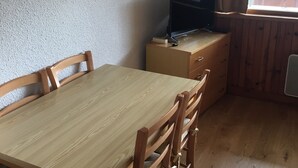 Ein kleines Zimmer mit einem Holztisch und Stühlen, einem Fernseher auf einer Kommode und einem Fenster mit Vorhängen.