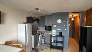 Eine kleine Küche mit Kühlschrank, Mikrowelle und Kaffeemaschine. Ein Esstisch mit Stühlen und ein kleiner Teppich sind vorhanden.