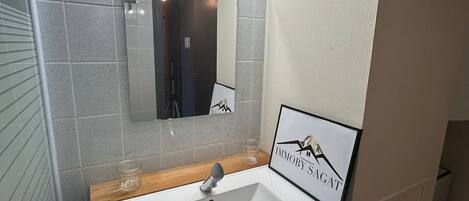 Une salle de bain avec un lavabo blanc, un miroir surmonté de lumières et un panneau encadré accroché au mur.