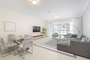 A modern living room with a glass dining table, grey sofas, a flat-screen TV, and a large window with curtains.