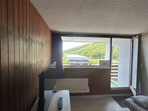 Une pièce aux murs en bois, avec un bureau doté d’un ordinateur et d’une télécommande, un radiateur et une porte-fenêtre coulissante donnant sur un balcon avec vue sur un immeuble et de la verdure.