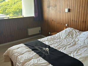 Une chambre avec un mur lambrissé en bois, un lit avec une literie blanche et une couverture noire, une fenêtre avec un rideau violet et une petite table de chevet.