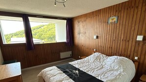 A bedroom with a large bed, wooden panel walls, a window with curtains, and a small table. - Central apartment, access to slopes, south balcony, WIFI, nearby parking (Besse-et-Saint-Anastaise-Super Besse)