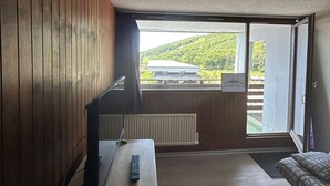 A room with wooden walls, a desk with a computer and a remote control, a radiator, and a sliding glass door leading to a balcony with a view of a building and greenery. - Central apartment, access to slopes, south balcony, WIFI, nearby parking (Besse-et-Saint-Anastaise-Super Besse)