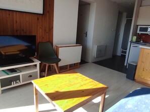 Quarto pequeno e compacto com televisão, uma cadeira, uma mesinha, cozinha compacta e porta para outro quarto.
