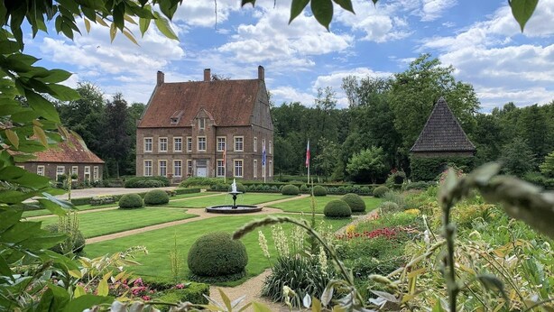 Ein großes, historisches Gebäude mit einer symmetrischen Fassade, einem formellen Garten mit einem Brunnen und einer Fahnenstange.